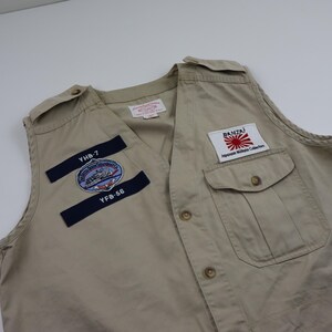 May include: A beige utility vest with multiple patches. The patches include a navy blue patch with the text "YHB-7" and "YFB-56", a circular patch with a ship and the text "United States Navy", and a white patch with the text "BANZAI" and a Japanese flag.