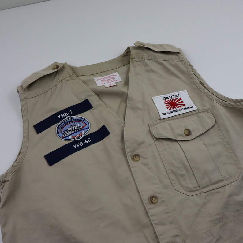 May include: A beige utility vest with multiple patches. The patches include a navy blue patch with the text "YHB-7" and "YFB-56", a circular patch with a ship and the text "United States Navy", and a white patch with the text "BANZAI" and a Japanese flag.