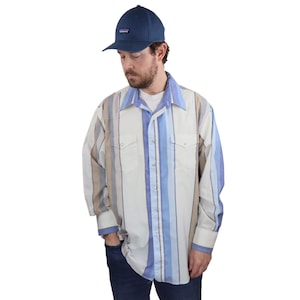 Puede incluir: Una camisa de manga larga a rayas con botones, fondo blanco y rayas azules y marrones. La camisa tiene un cuello puntiagudo y dos bolsillos en el pecho. La persona lleva pantalones vaqueros azules y una gorra de béisbol azul marino con un pequeño logotipo.