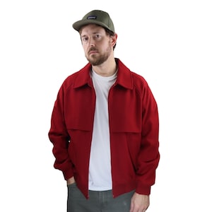 Könnte beinhalten: Eine rote Jacke mit durchgehendem Reißverschluss, Kragen und geripptem Saum. Die Jacke wird über einem weißen T-Shirt getragen und mit einer grünen Hose kombiniert. Auf dem Kopf wird eine grüne Baseballkappe getragen.