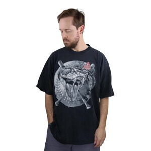 Może przedstawiać: Czarny t-shirt z szczegółową grafiką węża z otwartą paszczą, kijami do baseballu i piłką do baseballu. Projekt jest w odcieniach szarości i bieli, z odrobiną czerwieni. Koszulka jest noszona przez osobę.
