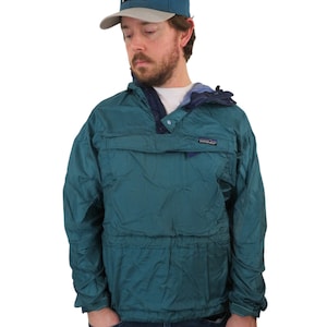 May include: Teal anorak jacket with a front pocket and a blue and gray baseball cap. The jacket has a drawstring at the waist and a small logo patch. The cap features a mountain logo.