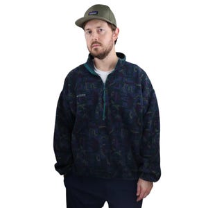 Puede incluir: Un jersey de polar azul oscuro con un patrón abstracto verde y morado. La cremallera está parcialmente cerrada y el cuello está levantado. El polar tiene el logotipo de Columbia en el pecho izquierdo. La persona lleva una gorra de béisbol verde con un pequeño parche en la parte delantera.