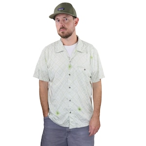 May include: A light green patterned shirt with a button-down front, worn with grey shorts and a green baseball cap. The shirt features a geometric design. The outfit is casual and suitable for everyday wear.