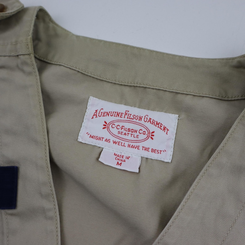 May include: A beige cotton shirt with a label that reads "AGENUINE FILSON GARMENT" and "CC-FILSON CO. SEATTLE" with the tagline "MIGHT AS WELL HAVE THE BEST". The label also states "MADE IN CHINA" and "M".