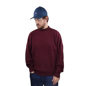 May include: A maroon mock neck sweatshirt with long sleeves and a relaxed fit. The sweatshirt has a subtle seam detail across the chest. The person is wearing a dark blue baseball cap with a small logo.