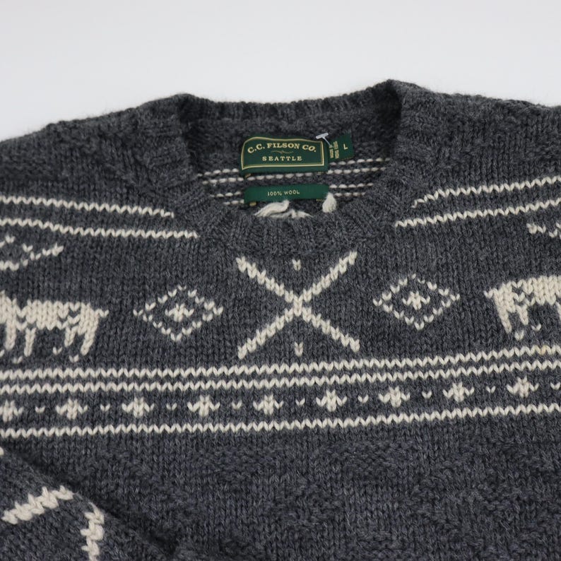 May include: A gray wool sweater with a white and gray Fair Isle pattern featuring reindeer and a cross design. The sweater has a crew neck and a label that reads "C.C. Filson Co. Seattle 100% Wool".