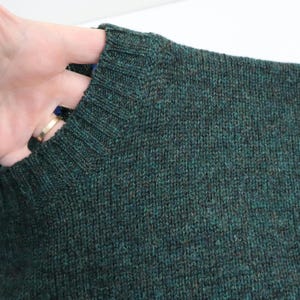 Pu&ograve; includere: Primo piano di un maglione lavorato a maglia verde scuro. Il maglione ha uno scollo a coste e un motivo a maglia strutturato. Il colore &egrave; un mix di verde scuro e marrone, creando un effetto m&eacute;lange. La manica &egrave; visibile, tenuta da una mano.