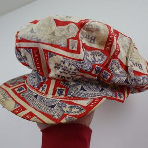 以下が含まれることがあります： 赤、白、青の模様にテキストとロゴが入ったヴィンテージスタイルのキャップ。 つばと丸みを帯びたクラウンが付いています。 デザインには、繰り返しのパターンで「GENUINE」と「Busch」という言葉が含まれています。