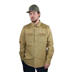 Puede incluir: Una camisa color beige con parches de ante en los hombros y detalles en los bolsillos. La camisa tiene dos bolsillos en el pecho y se combina con una gorra de béisbol verde. El conjunto es informal y adecuado para actividades al aire libre.