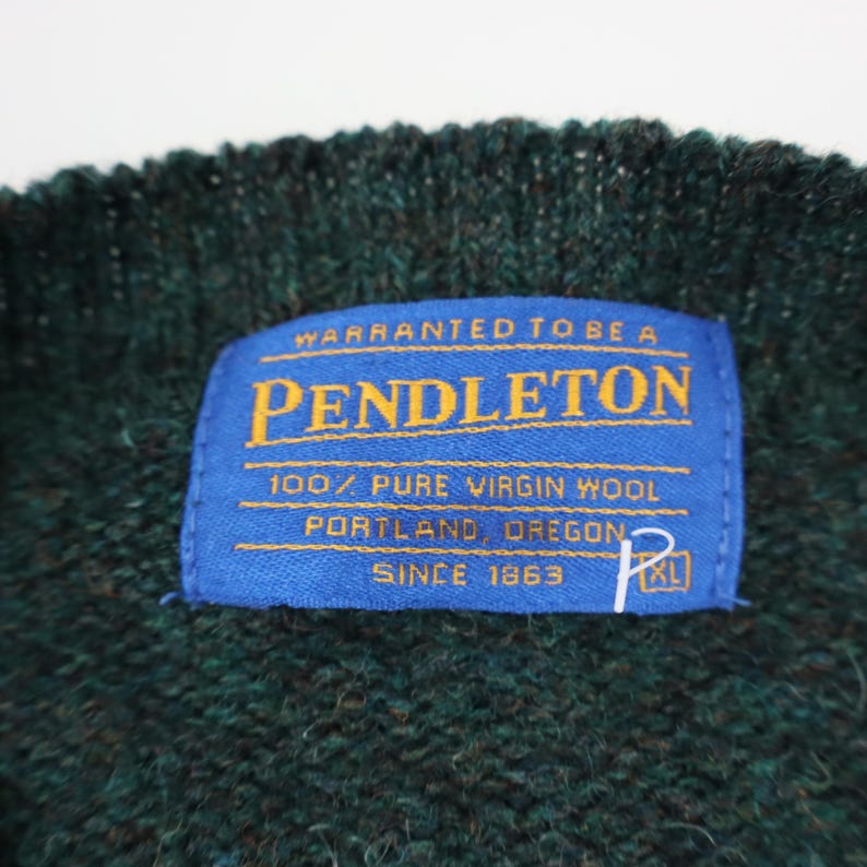 Pu&ograve; includere: Primo piano di un maglione lavorato a maglia verde scuro con un'etichetta blu. L'etichetta recita "PENDLETON" e "100% PURE VIRGIN WOOL, PORTLAND OREGON, SINCE 1863". Il maglione &egrave; XL.
