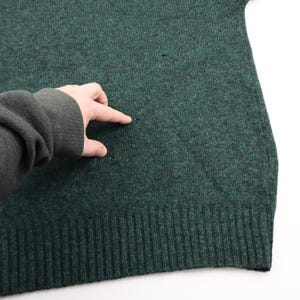 Pu&ograve; includere: Primo piano di un maglione verde scuro con orlo e polsini a coste. Il maglione presenta alcuni piccoli fori. La mano di una persona indica uno dei fori.