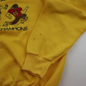 Puede incluir: Sudadera amarilla con un gr&aacute;fico de jugadores de f&uacute;tbol y el texto "CHAMPIONS".