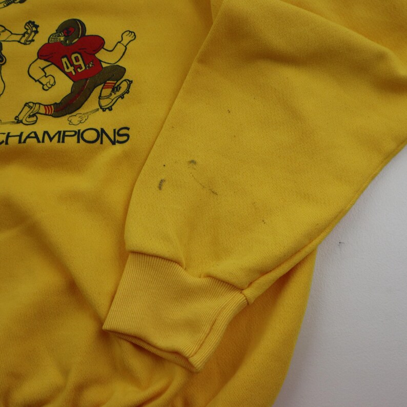 Puede incluir: Sudadera amarilla con un gr&aacute;fico de jugadores de f&uacute;tbol y el texto "CHAMPIONS".