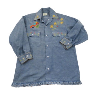 Puede incluir: Una camisa de mezclilla azul claro con flores bordadas y un pájaro en la parte delantera. La camisa tiene un dobladillo con volantes y mangas largas con puños bordados.