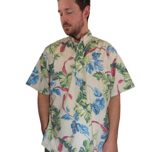 以下が含まれることがあります： クリーム色のボタンダウンの半袖シャツで、緑、青、赤の色の熱帯の花柄がプリントされています。シャツの右胸には「NHYC」と刺繍された青いロゴがあります。
