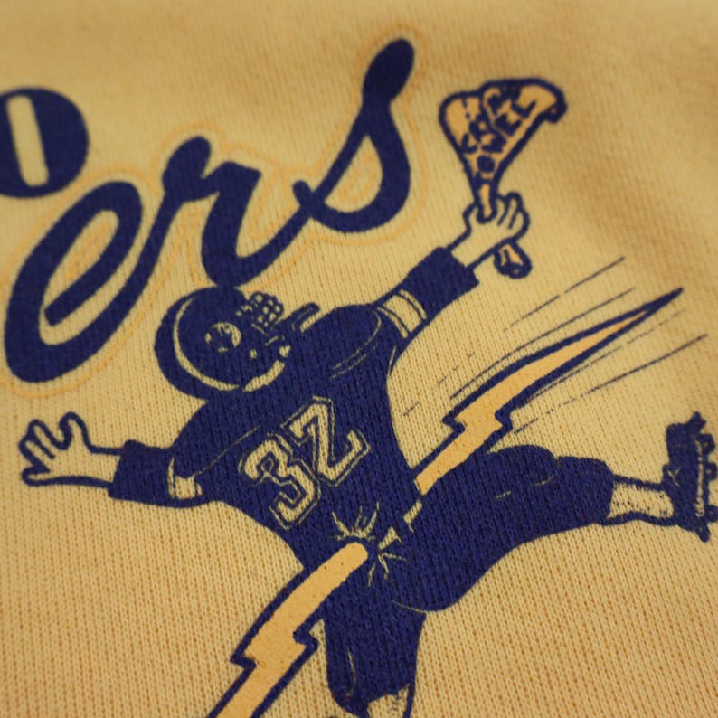 Puede incluir: Una sudadera amarilla con un gr&aacute;fico azul de un jugador de f&uacute;tbol americano corriendo con un rayo detr&aacute;s de &eacute;l. El jugador lleva una camiseta azul con el n&uacute;mero 32. El texto "Goers" est&aacute; escrito en azul sobre el jugador.