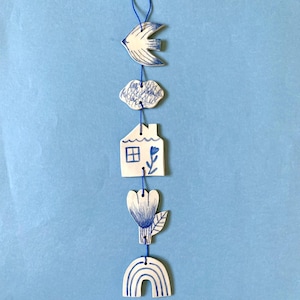 Peut inclure: Une suspension murale en céramique blanche avec des détails peints en bleu. La suspension représente un oiseau, un nuage, une maison, une fleur et un arc-en-ciel.