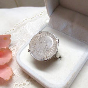 Puede incluir: Un anillo de plata con una cara grande y redonda en forma de moneda, engastado en una sencilla banda de plata. El anillo se muestra en una caja de joyería blanca. Una rosa rosa es visible en el fondo.