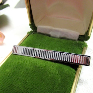 May include: A silver-toned metal tie bar featuring a repeating vertical pinstripe pattern across its rectangular surface. The tie bar is displayed resting inside an open, hinged jewelry box lined with vibrant green velvet fabric.