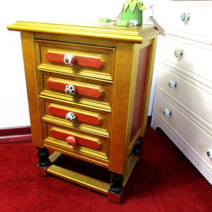 Könnte beinhalten: Ein kleiner Nachttisch aus Holz mit vier Schubladen. Der Nachttisch ist goldfarben lackiert mit roten Akzenten und schwarzen Füßen. Jede Schublade hat einen einzigartigen, dekorativen Knopf. Auf der Oberseite sitzt eine grüne Figur.