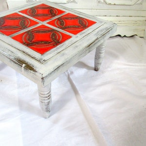 Könnte beinhalten: Ein quadratischer Beistelltisch mit vier gedrehten Beinen, weiß lackiert. Die Tischplatte ist mit vier roten und goldenen Fliesen mit geometrischem Muster verziert.
