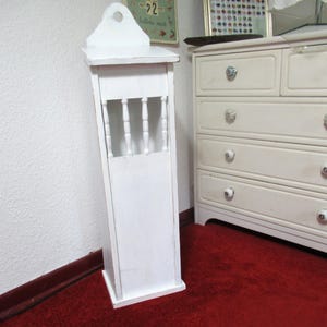 Könnte beinhalten: Ein weißer Holzschrank mit dekorativer Oberseite und einer kleinen Öffnung mit vertikalen Spindeln. Der Schrank ist weiß lackiert und steht auf einem roten Teppich. Eine weiße Kommode befindet sich im Hintergrund.