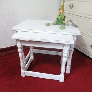 Könnte beinhalten: Ein Satz weißer Beistelltische mit gedrechselten Beinen. Eine kleine Holzfigur mit grünem Outfit und Hut sitzt obenauf. Die Tische stehen auf einem roten Teppich. Im Hintergrund ist eine weiße Kommode. Die Tische sind ca. 40 cm lang.