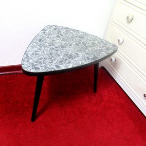 Könnte beinhalten: Kleiner, dreieckiger Beistelltisch mit einer gesprenkelten, grauen Tischplatte und schwarzen Beinen. Der Tisch steht auf einem roten Teppich, im Hintergrund eine weiße Kommode. Maße: ca. 61 cm breit und 46 cm tief.
