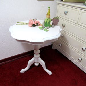 Könnte beinhalten: Kleiner, weißer Tisch mit gewelltem Rand, Säulenfuß und drei geschwungenen Beinen. Auf dem Tisch liegen ein Strauß rosa Blumen und eine Holzfigur. Im Hintergrund eine weiße Kommode mit dekorativen Knöpfen.