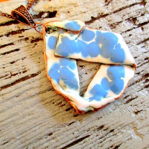 Op de afbeelding: Een blauwe en witte keramische hanger met een bloemenpatroon hangt aan een koperen ketting. De hanger heeft een onregelmatige vorm en heeft een rustieke, handgemaakte uitstraling.