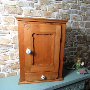 Könnte beinhalten: Ein hellbrauner Wand-Holzschrank mit einer einzelnen Tür mit dekorativem Schnitzpaneel und einer kleinen unteren Schublade. Sowohl die Tür als auch die Schublade sind mit runden, weißen Keramikknöpfen ausgestattet, die mit kleinen blauen Blumenmustern verziert sind.