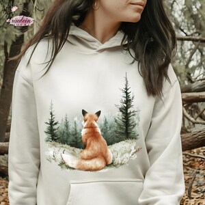 Könnte beinhalten: Ein cremefarbener Kapuzenpullover mit einer Aquarellillustration eines Fuchses, der in einem Wald sitzt. Der Fuchs hat orangefarbenes Fell und ist vom Betrachter abgewandt. Die Waldszene umfasst grüne Bäume und einen nebligen Hintergrund. Der Hoodie hat eine Kapuze mit Kordelzug und eine Vordertasche.