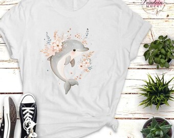 Könnte beinhalten: Weißes T-Shirt mit einer Aquarellillustration eines grauen Delfins, umgeben von Blumen und Blättern. Der Delfin hat einen lächelnden Ausdruck. Der Text "Fräulein Mucki" befindet sich oben rechts. Schwarz-weiße Turnschuhe sind unten links.