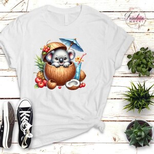 Könnte beinhalten: Weißes T-Shirt mit einem Cartoon-Koala in einer Kokosnussschale, mit einem blauen Sonnenschirm und einem tropischen Getränk. Das Design umfasst Kirschen, Ananas und eine halbe Kokosnuss. Schwarz-weiße Turnschuhe sind unten zu sehen.