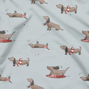 Könnte beinhalten: Hellblauer Stoff mit einem sich wiederholenden Muster von Cartoon-Dackeln. Einige Hunde tragen rot-weiß gestreifte Badehosen, andere haben rot-weiß gestreifte Schwimmreifen, und einige sind auf Surfbrettern. Der Hintergrund hat kleine schwarze Sprenkel.