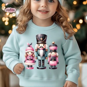 Könnte beinhalten: Hellblaues Sweatshirt mit einem Aufdruck von drei Nussknackern in Rosa, Schwarz und Türkis. Die Nussknacker haben detaillierte Uniformen und Hüte. Das Sweatshirt wird von einem Kind getragen.