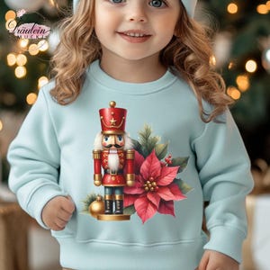 Könnte beinhalten: Hellblaues Sweatshirt mit farbenfrohem Weihnachtsdesign. Das Design umfasst einen rot-goldenen Nussknacker, einen roten Weihnachtsstern und eine goldene Kugel. Das Sweatshirt hat den Text "Fräulein Mucki" in der oberen linken Ecke.