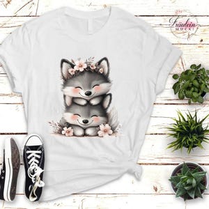 Könnte beinhalten: Weißes T-Shirt mit einer gestapelten Illustration von zwei lächelnden grauen Wolfsjungen mit Blumenkränzen und rosigen Wangen. Das Design beinhaltet rosa Blumen. Der Hintergrund ist weißes Holz. Schwarz-weiße Turnschuhe sind sichtbar.