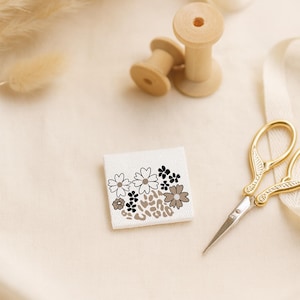 Könnte beinhalten: Ein kleines, quadratisches weißes Stoffetikett mit einem floralen Design in Schwarz, Grau und Braun. Das Etikett befindet sich neben einer goldenen Schere und Holzspulen. Der Hintergrund ist ein neutrales Beige.
