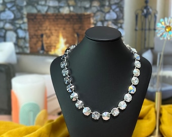 May include: A silver necklace with clear crystal beads on a black display stand. The necklace is in front of a fireplace with a yellow blanket.