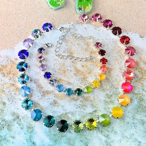 May include: A rainbow-coloured crystal necklace with a silver chain. The necklace is made of round, faceted crystals in shades of blue, green, yellow, orange, and red. The necklace is laid out on a sandy surface.