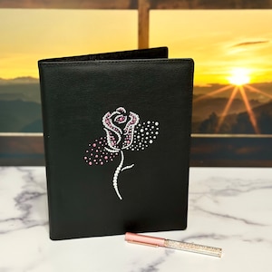 Könnte beinhalten: Ein schwarzes Lederjournal mit einem Rosenmotiv aus rosa und weißen Strasssteinen. Das Journal ist geschlossen und liegt auf einer weißen Marmorfläche. Ein roségoldener Stift mit Strasssteinen liegt neben dem Journal.