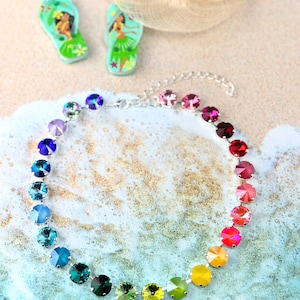 May include: A rainbow-coloured crystal necklace with a silver chain and round, faceted stones. The necklace is on a sandy background with a blue and white wave pattern.