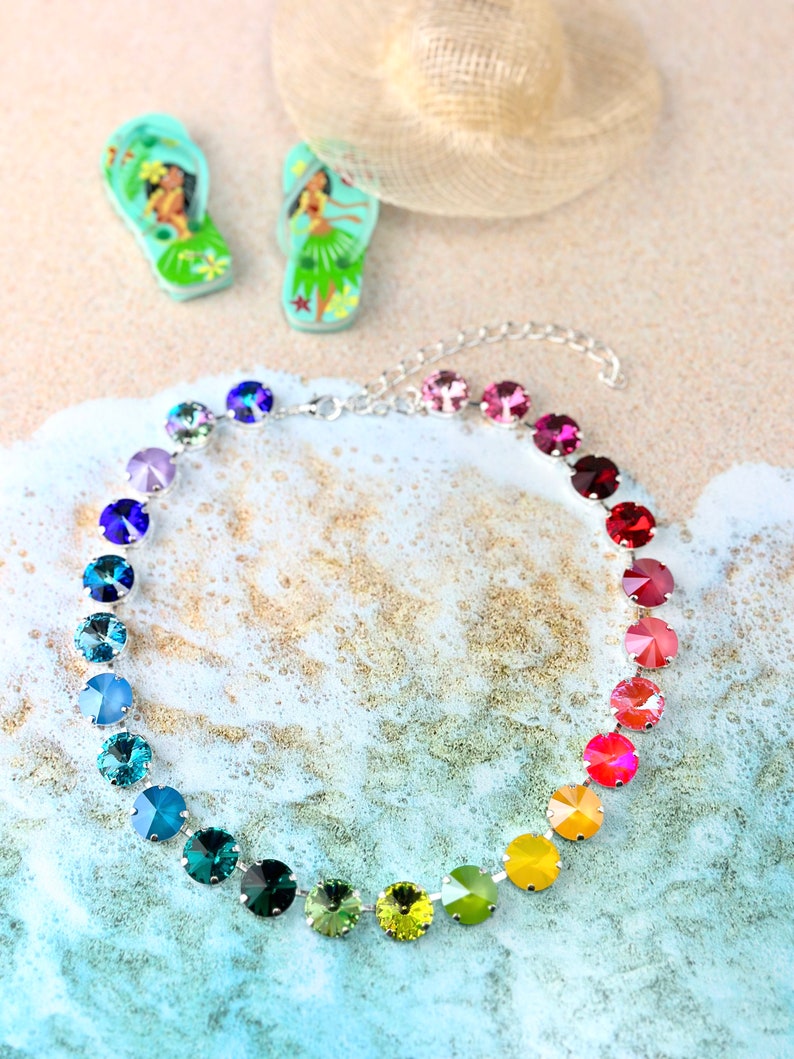 May include: A rainbow-coloured crystal necklace with a silver chain and round, faceted stones. The necklace is on a sandy background with a blue and white wave pattern.
