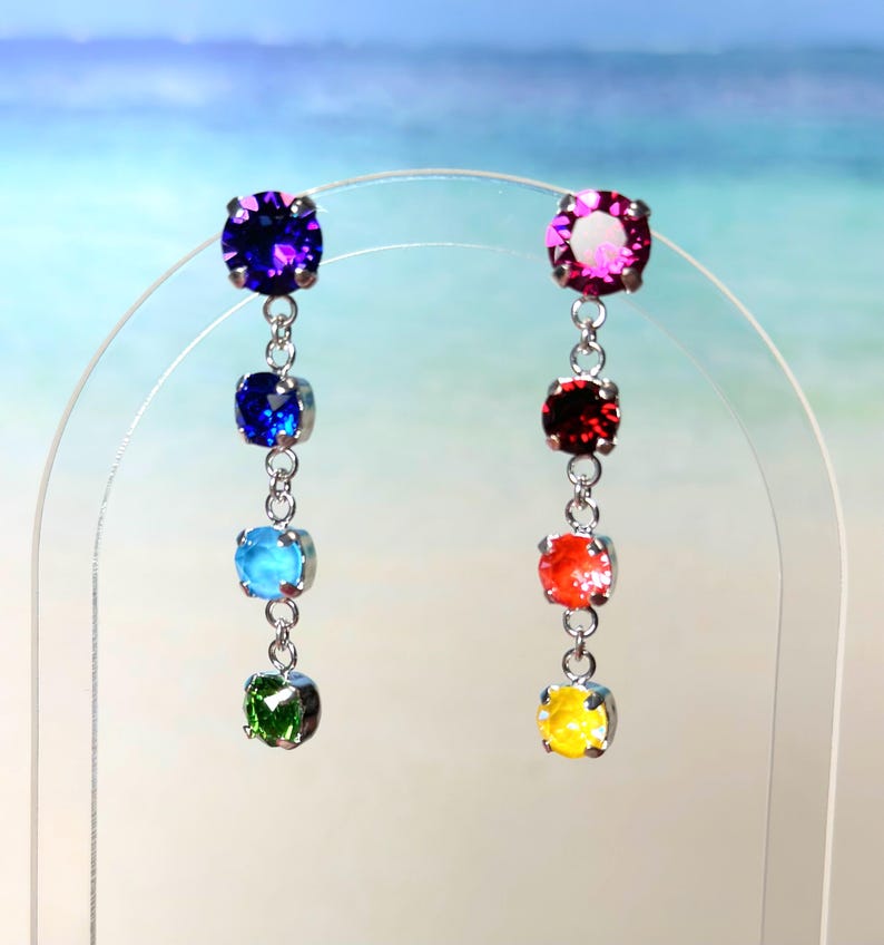 May include: A pair of dangle earrings featuring a rainbow of colours. The earrings are made with silver chains and round, faceted gemstones in shades of purple, blue, turquoise, green, orange, and yellow.