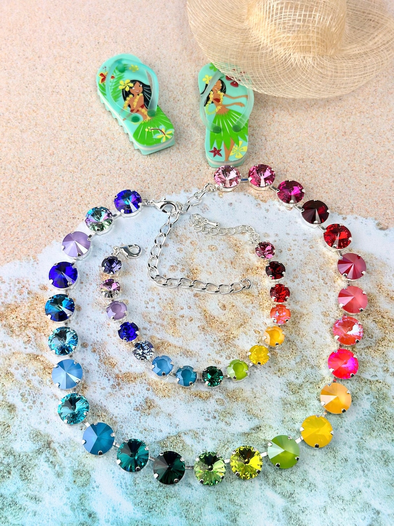 May include: A rainbow-coloured crystal necklace with a silver chain. The necklace is made of round, faceted crystals in shades of blue, green, yellow, orange, and red. The necklace is laid out on a sandy surface.