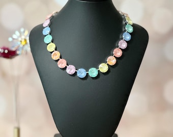 May include: A colourful statement necklace on a black mannequin. The necklace has large, round, faceted beads in pastel shades of pink, yellow, blue, and green. The necklace is displayed on a wooden stand with a blurred background.