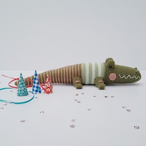 May include: A handmade, green and brown crochet alligator toy with a striped belly and a smiling face. The alligator is lying down next to three party hats and colorful ribbons. A fun, whimsical toy.