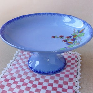 May include: A light blue ceramic pedestal cake stand with a wide, shallow bowl and a rounded base. The rim of the bowl has a darker blue, textured edge. Floral and leaf designs in red, orange, and green decorate the side.